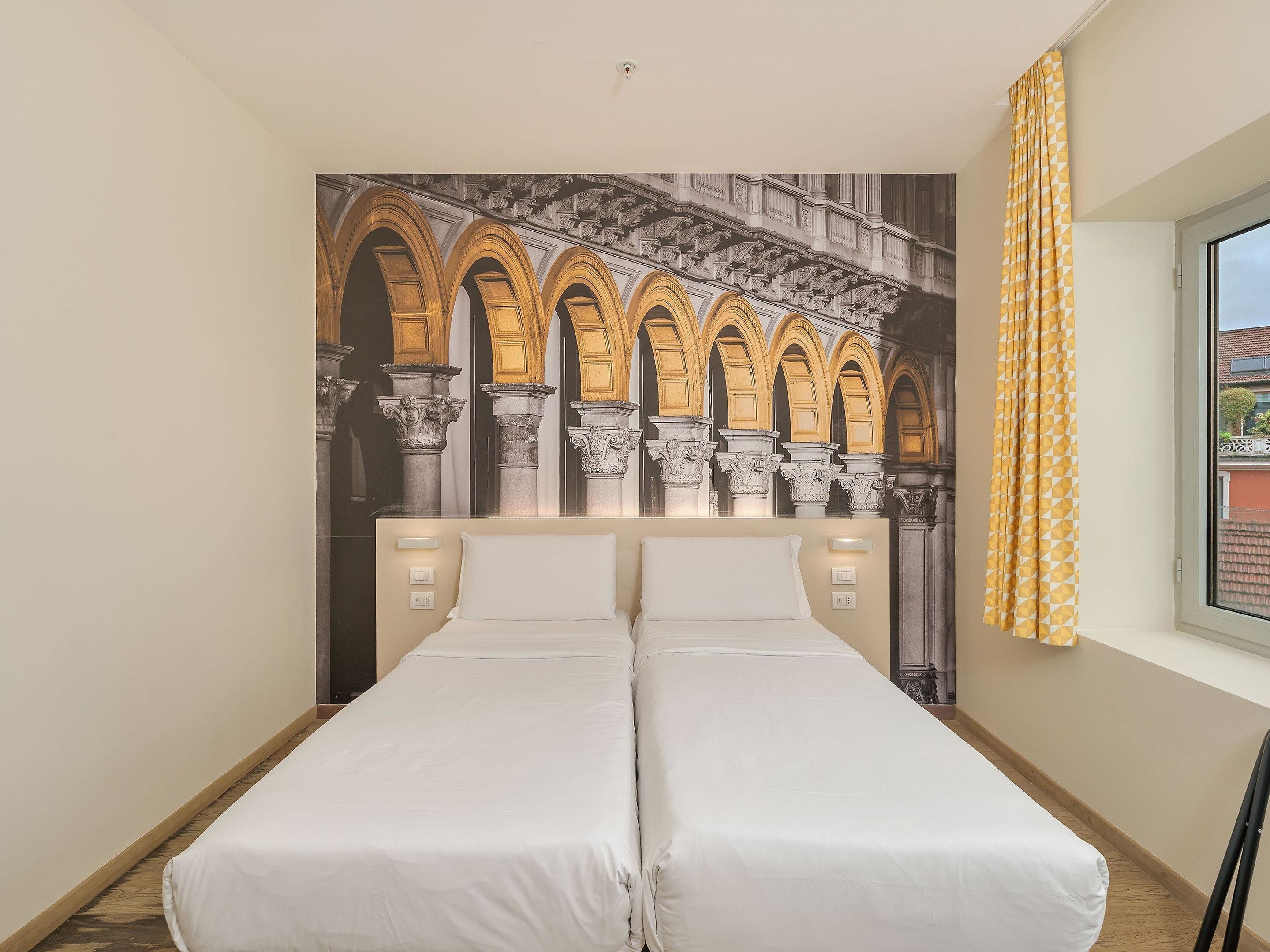 Ein Zimmer mit zwei Betten, einem Wandgemälde von Bögen, einem Fenster mit Vorhängen und Blick auf Gebäude.
