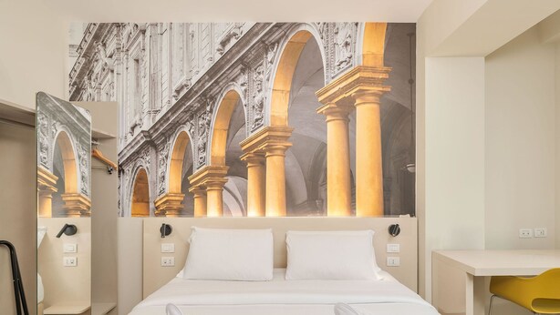 A hotel room with a large bed, two white pillows, a desk, a chair, and a large mural of an architectural scene. - B&B Hotel Milano Central Station (Milan)