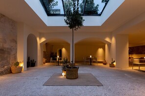 Relax in a spacious interior with a central column, seating, and tables, surrounded by lush greenery and warm ambiance.