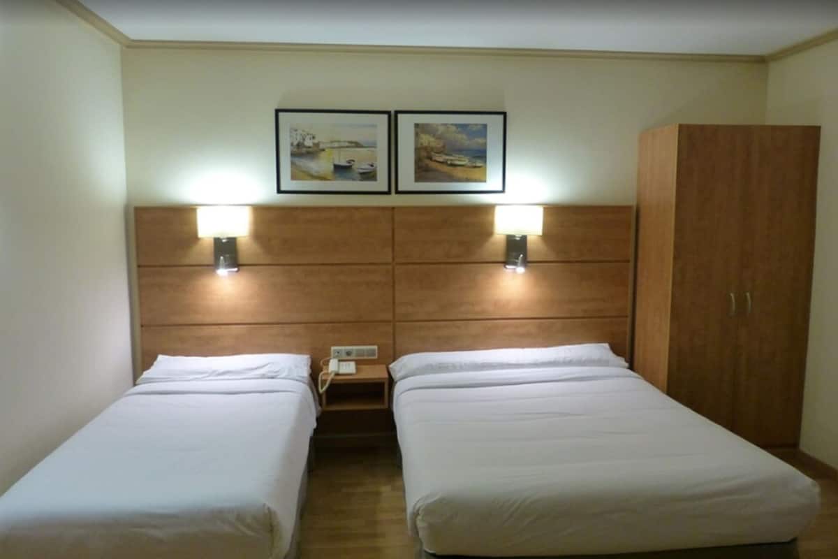 Una habitación de hotel con dos camas individuales, cabecero de madera, dos lámparas de pared, una mesita de noche con teléfono y un armario de madera.