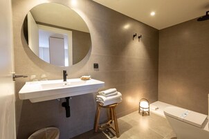 Een moderne badkamer met een witte wastafel, een ronde spiegel, een toilet en een klein krukje met handdoeken.