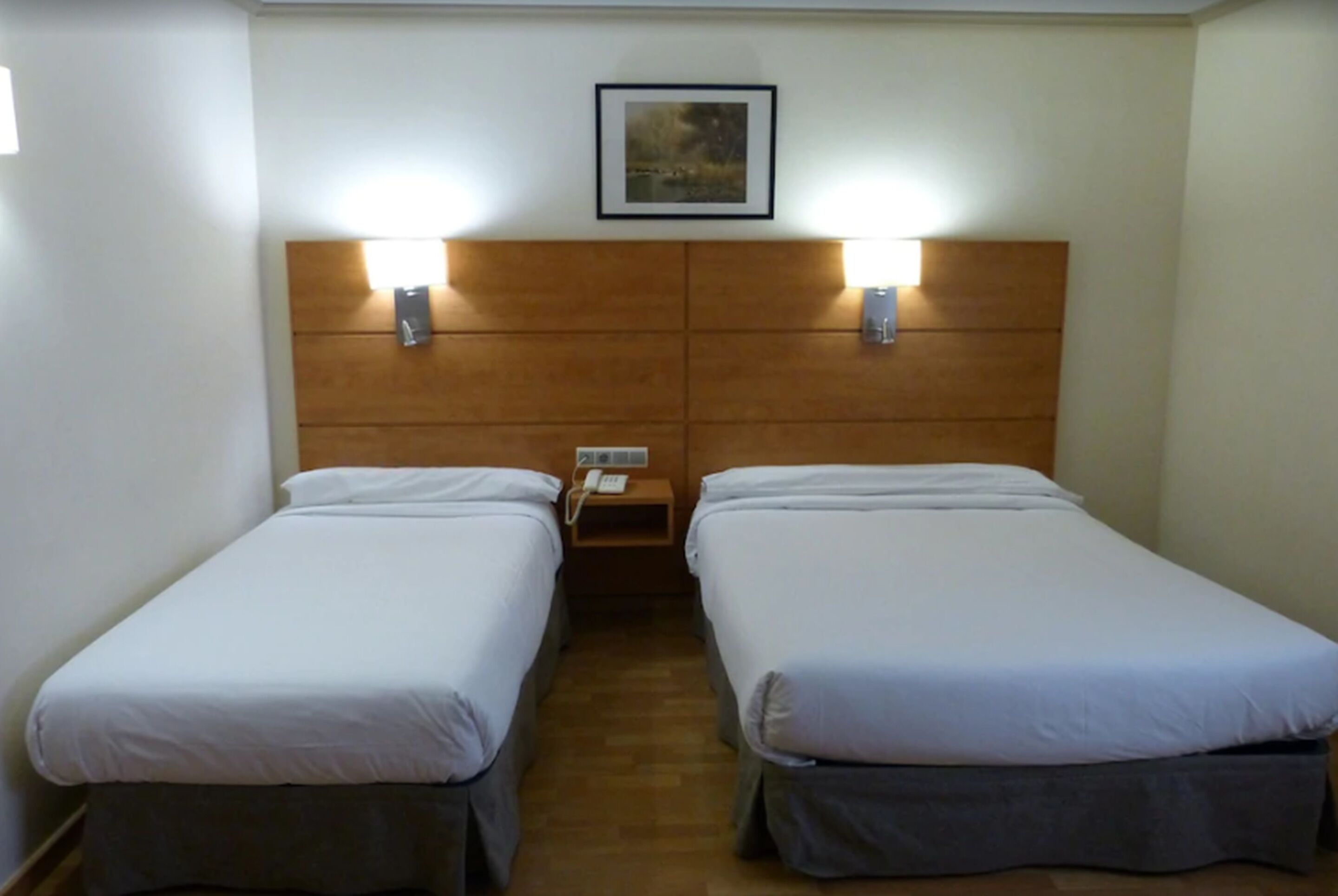 A hotel room with two single beds, a wooden headboard, bedside lamps, a small bedside table with a phone, and a framed picture on the wall.