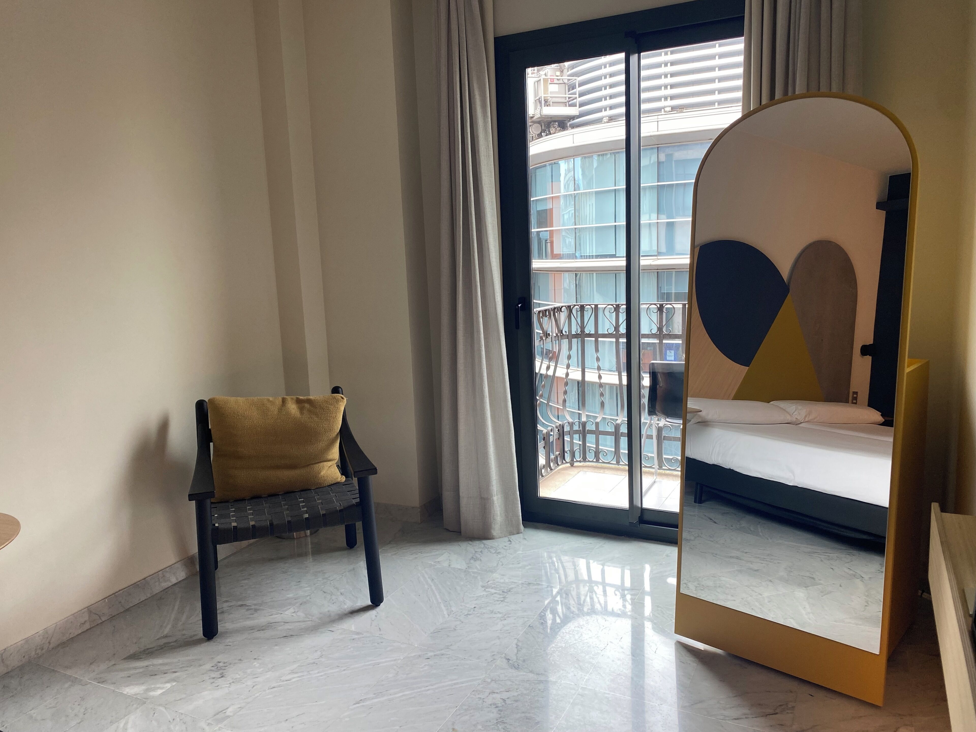 Habitación de hotel con cama, una silla, un espejo grande y balcón con vistas a edificios.
