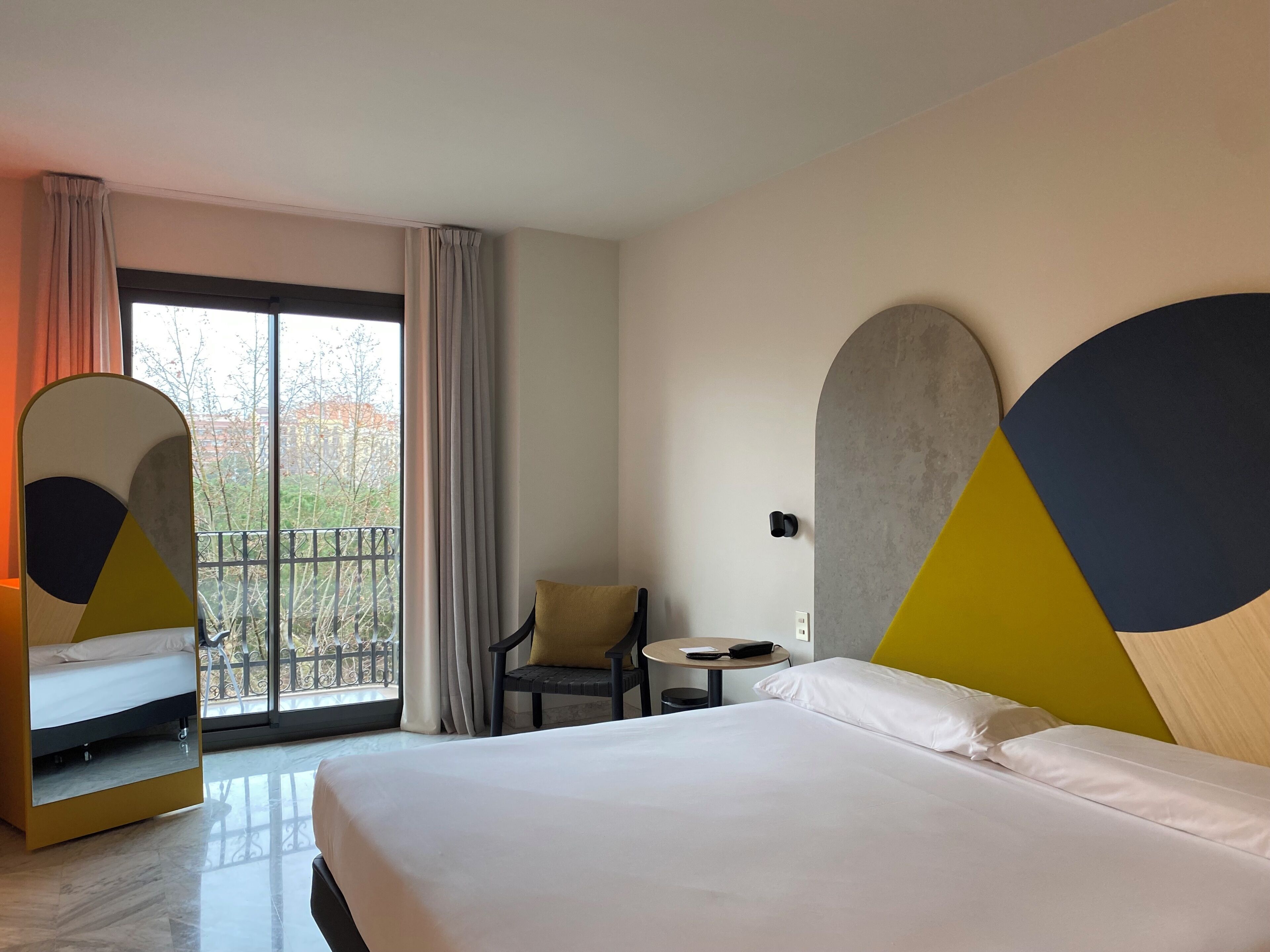 A hotel room with a bed, a chair, a small table, and a balcony with curtains.