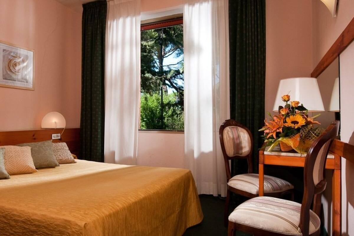 A hotel room with a bed, a desk with a lamp, a chair, and a window with curtains.