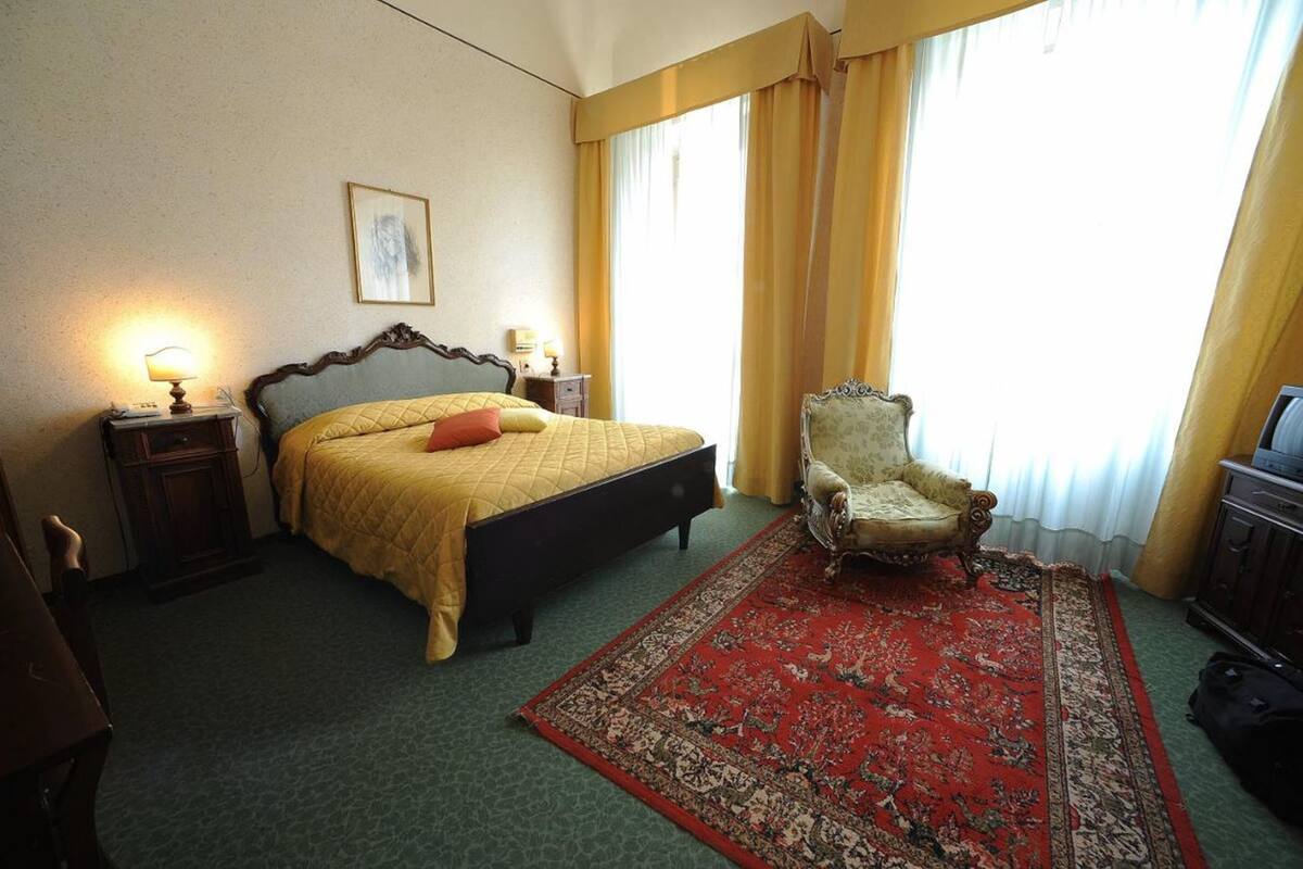 A hotel room with a bed, a chair, a nightstand, and a large window with curtains.