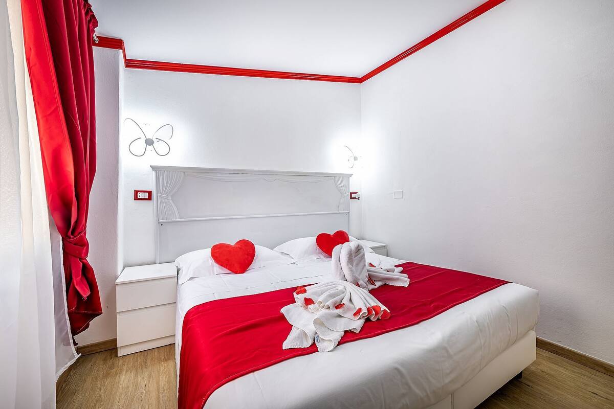 Una cama bien hecha con ropa de cama blanca y detalles rojos, una mesita de noche blanca y una cortina roja.