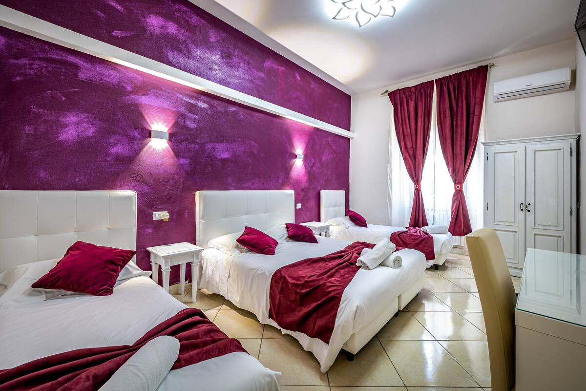 Habitación de hotel con dos camas, un armario blanco, una mesa de vidrio y una pared de acento morada.