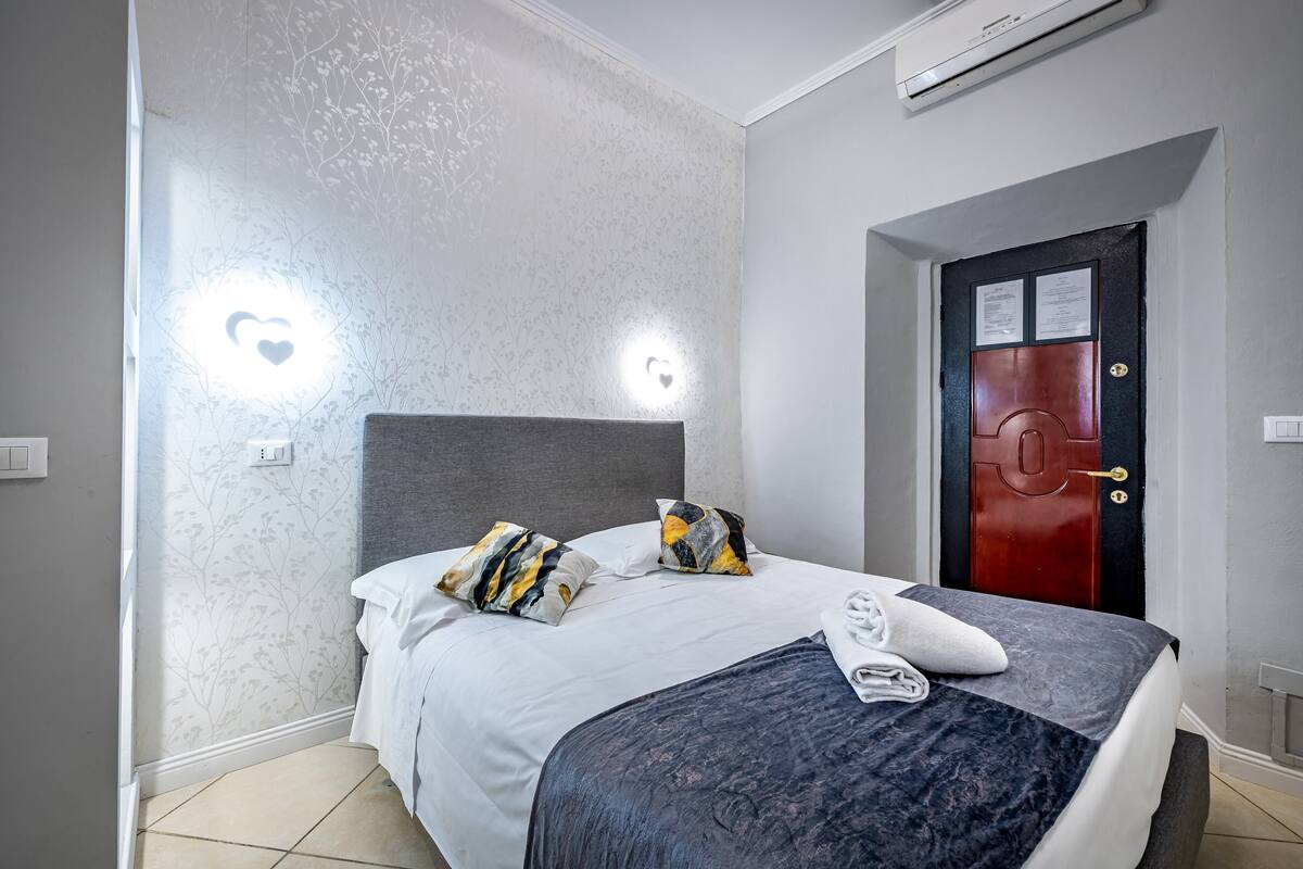 Una cama bien hecha con cabecera gris, sábanas blancas y almohadas estampadas. Se ve una puerta roja con cerradura a la derecha.