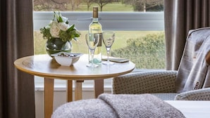 A hotel room with a view of a cityscape through large windows, a round wooden table with a bottle of wine, glasses, and a bowl of chocolates. - Tewkesbury Park (Tewkesbury)