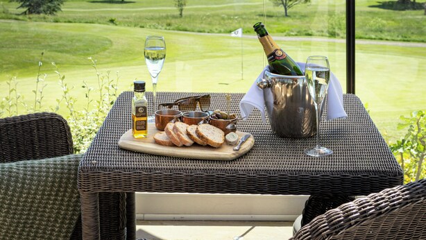 A table with a bottle of champagne, glasses, and a plate of food, set under a white umbrella with "Taittinger" written on it, overlooking a golf course. - Tewkesbury Park (Tewkesbury)
