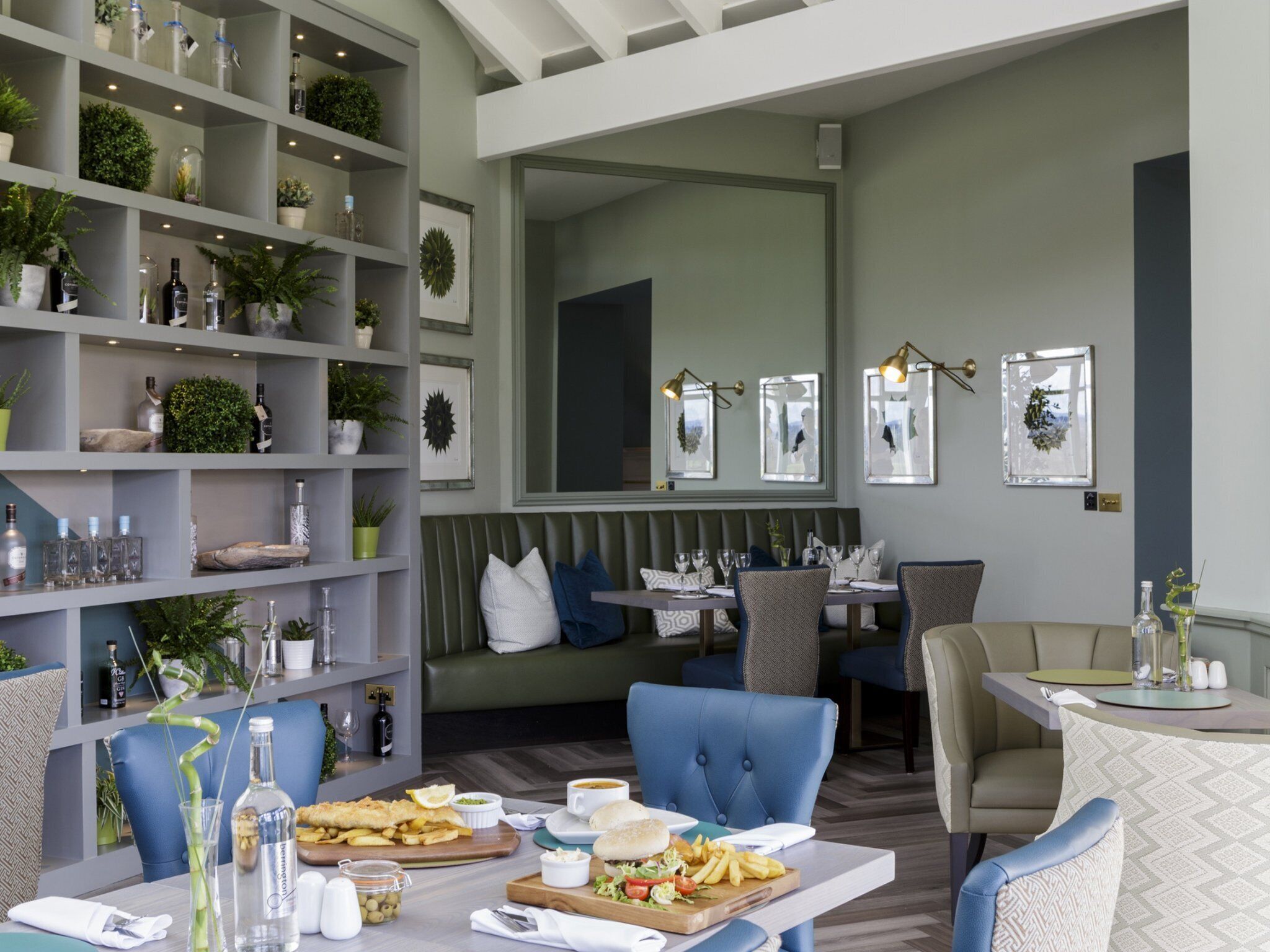 Ein Restaurant mit modernem Interieur, das eine lange Theke mit Sitzgelegenheiten, Regale mit Flaschen und Pflanzen sowie gerahmte Bilder an den Wänden bietet.