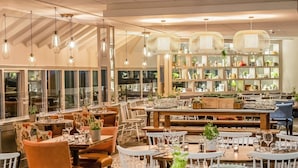 A restaurant with multiple dining tables and chairs, some with plants. There are hanging lights and a large window with a view. - Tewkesbury Park (Tewkesbury)