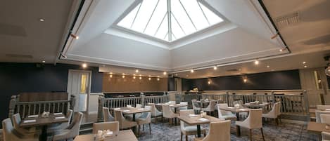 A spacious restaurant with a high ceiling and skylight, featuring multiple dining tables and chairs.