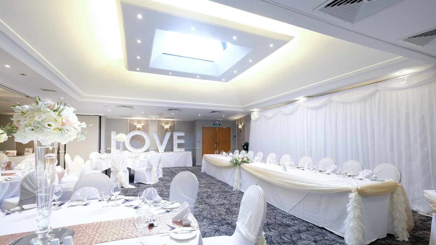 Un salón de banquetes con mesas redondas, manteles blancos y centros de mesa florales.