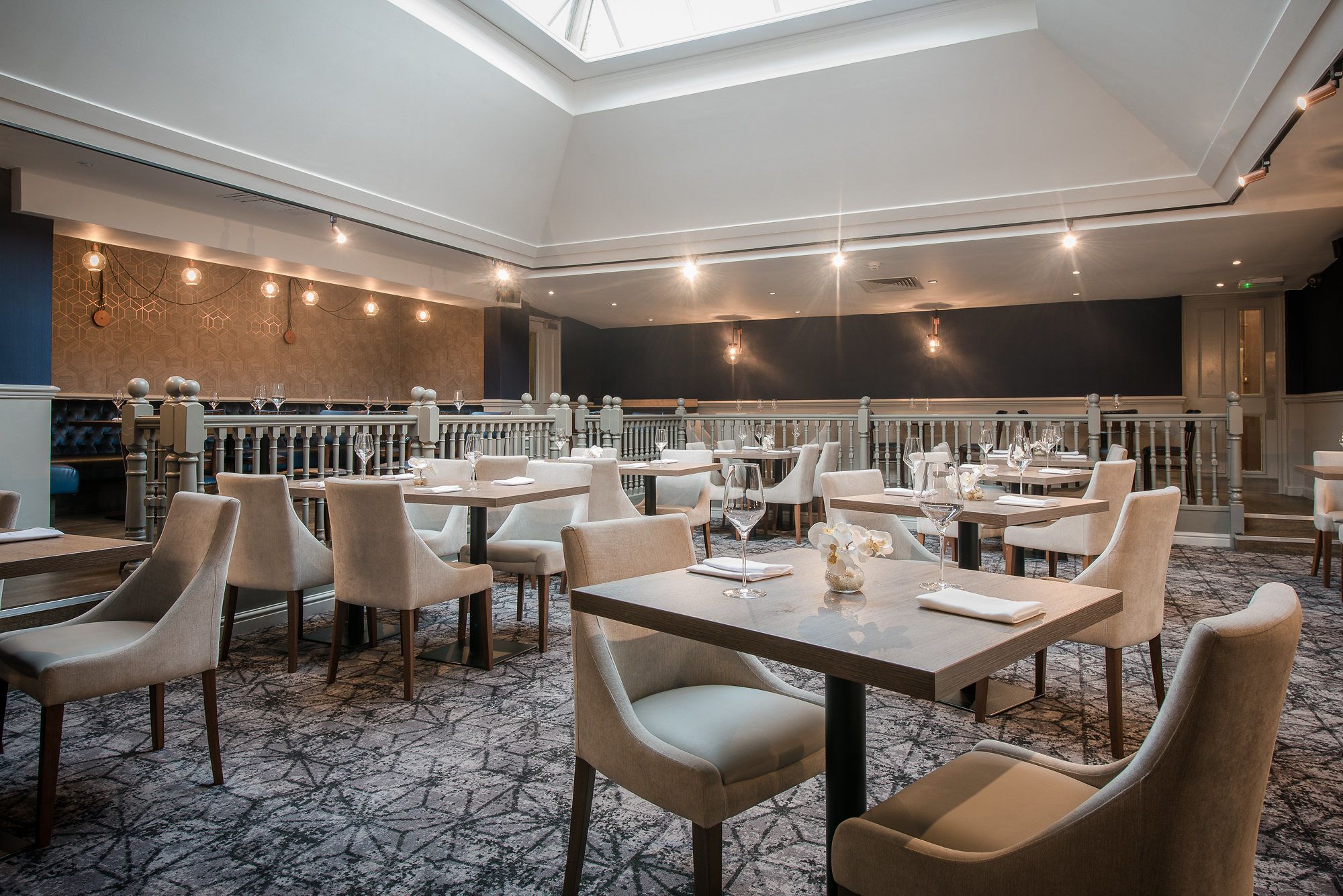 A modern restaurant with a high ceiling, geometric patterned carpet, and a variety of seating arrangements including tables and chairs.