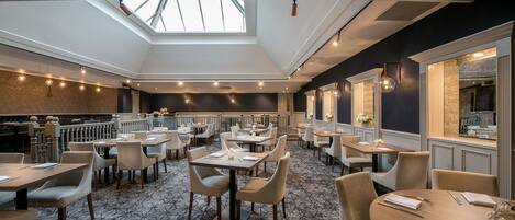 Un restaurant spacieux avec de hauts plafonds et des verrières, proposant un mélange de tables rondes et rectangulaires, ainsi qu’un tapis à motifs.