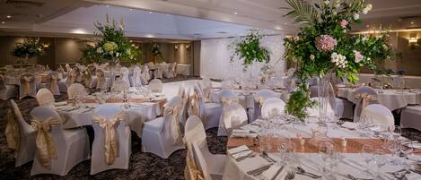 Une salle de banquet avec des tables rondes dressées pour un événement formel, chacune étant recouverte d’une nappe blanche, ornée d’un ruban doré et agrémentée d’un arrangement floral au centre.