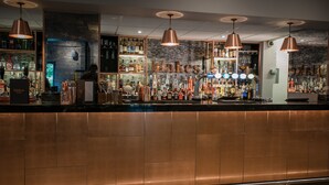 Zona de bar con mostrador cobrizo, lámparas colgantes de cobre y varias botellas y vasos en estantes.