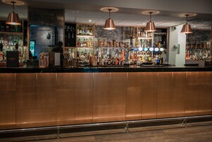 Zona de bar con mostrador cobrizo, lámparas colgantes de cobre y varias botellas y vasos en estantes.