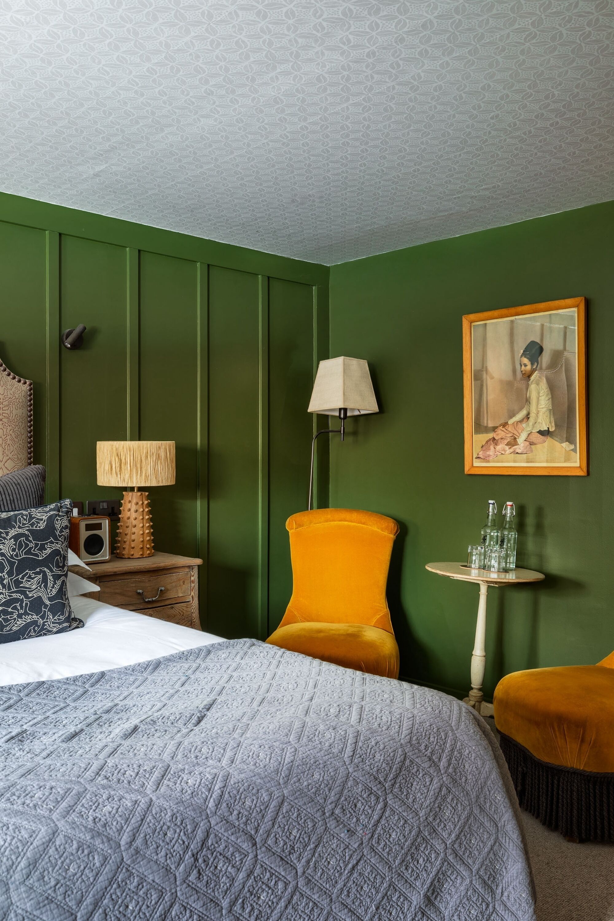 Een kamer met een bed, een nachtkastje met lamp, een stoel, een kleine tafel met flessen, en een lijst aan de muur.