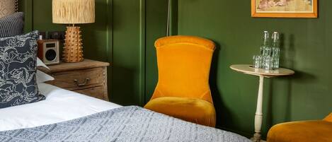 Een kamer met een bed, een nachtkastje met lamp, een stoel, een kleine tafel met flessen, en een lijst aan de muur.
