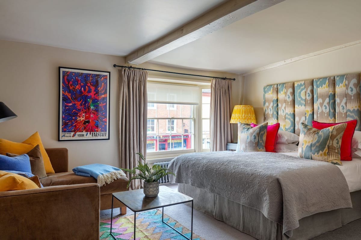 A hotel room with a bed, a chair, a small table with a plant, a colorful rug, and a large window with curtains.