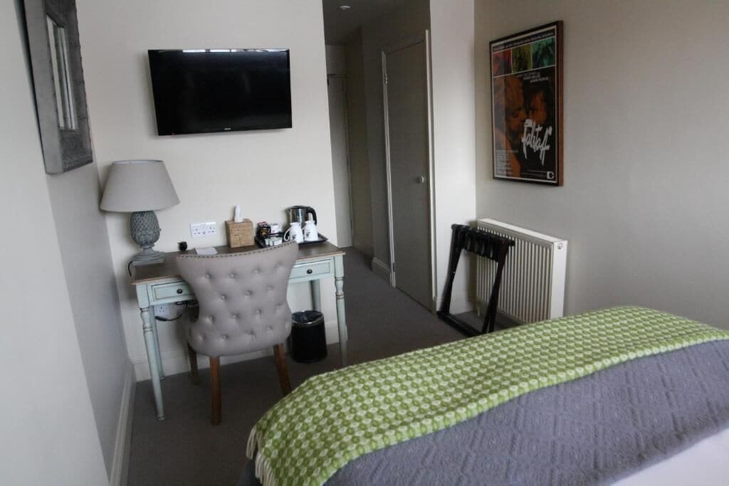 A hotel room with a bed, a desk with a chair, a television, a radiator, and a poster on the wall.