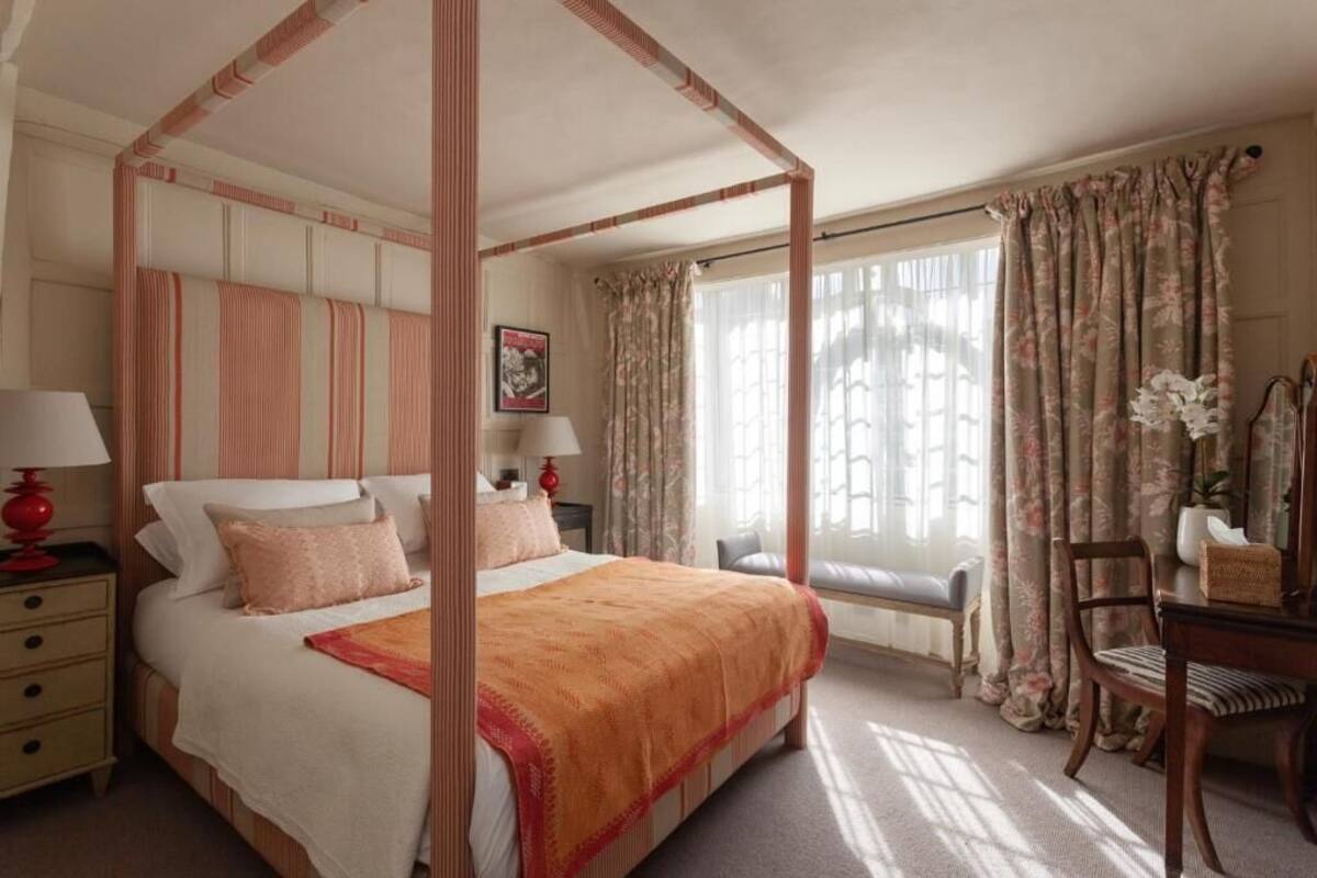 A four-poster bed with a canopy, a bedside table with a lamp, a chair, and a window with curtains.