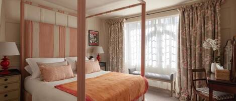 A four-poster bed with a canopy, a bedside table with a lamp, a chair, and a window with curtains.