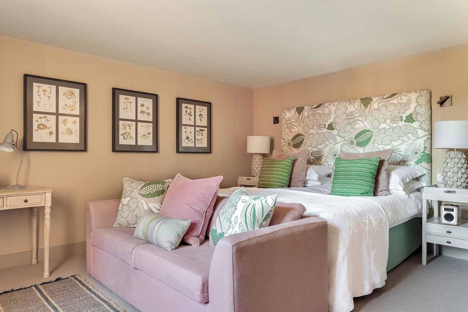 A bedroom with a bed, a sofa, a nightstand, a lamp, and framed botanical prints on the wall.