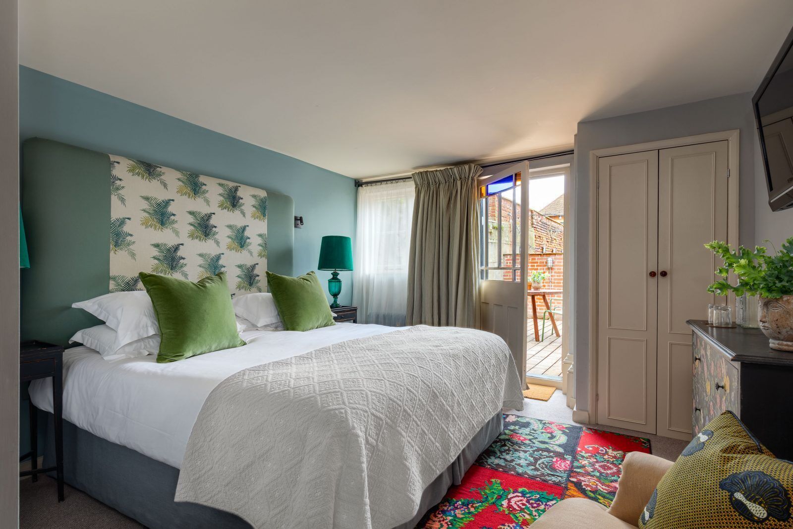 A bedroom with a bed, green pillows, a nightstand, a lamp, a dresser, and a view of the outside.