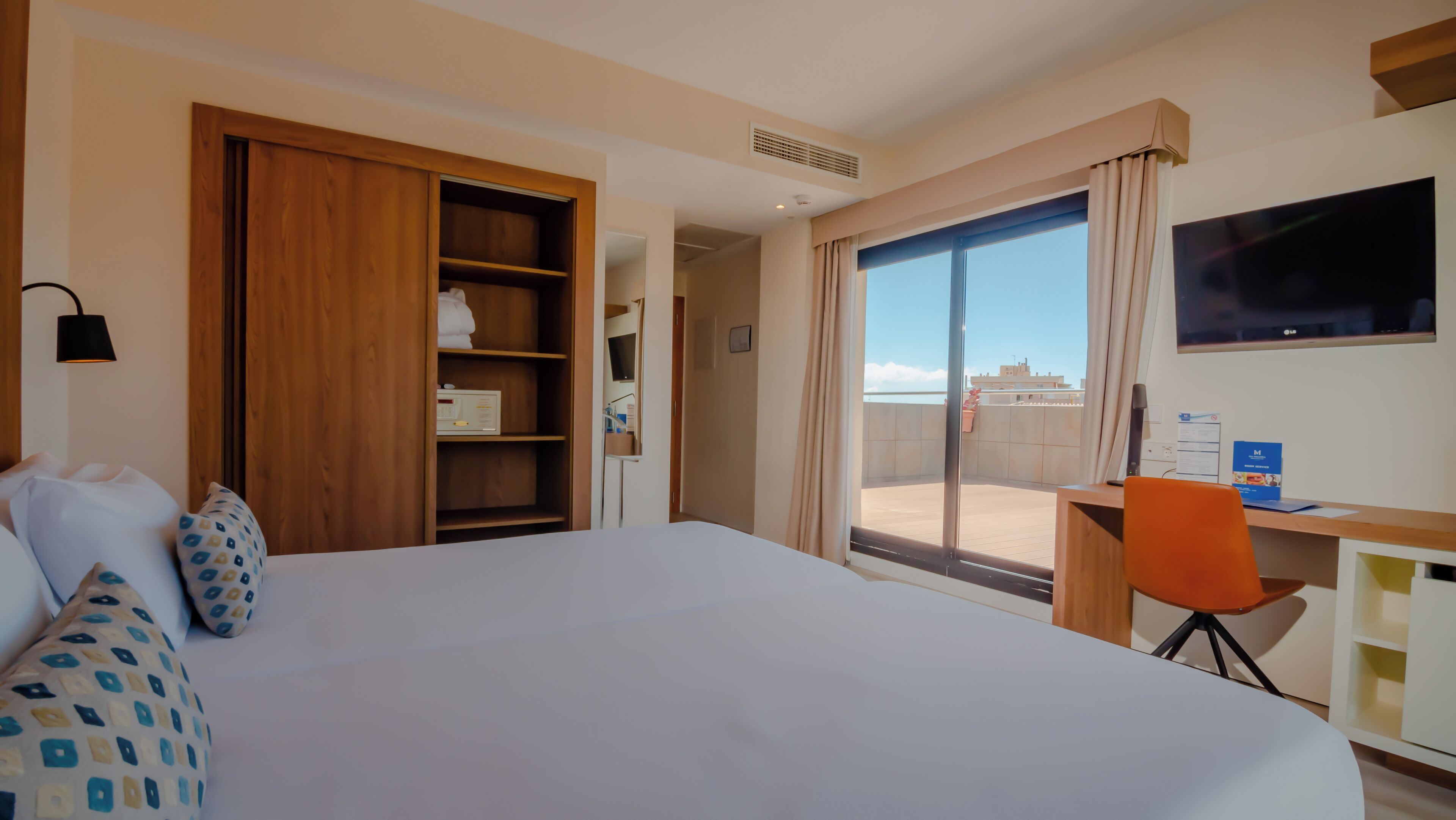 A hotel room with a large bed, a desk with a chair, a TV, and a balcony with a view of buildings.