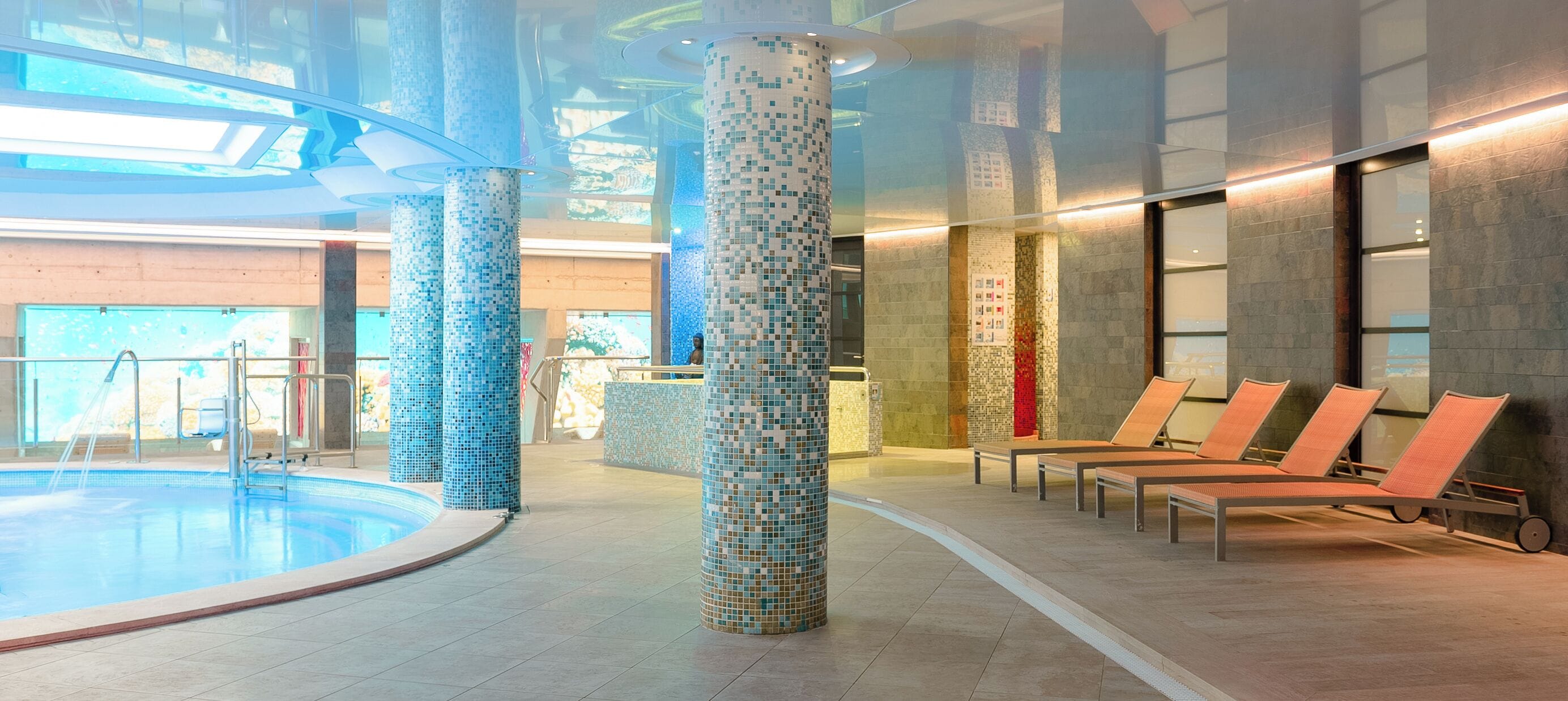 A modern indoor pool area with mosaic-tiled pillars, lounge chairs, and a clear view of the pool.