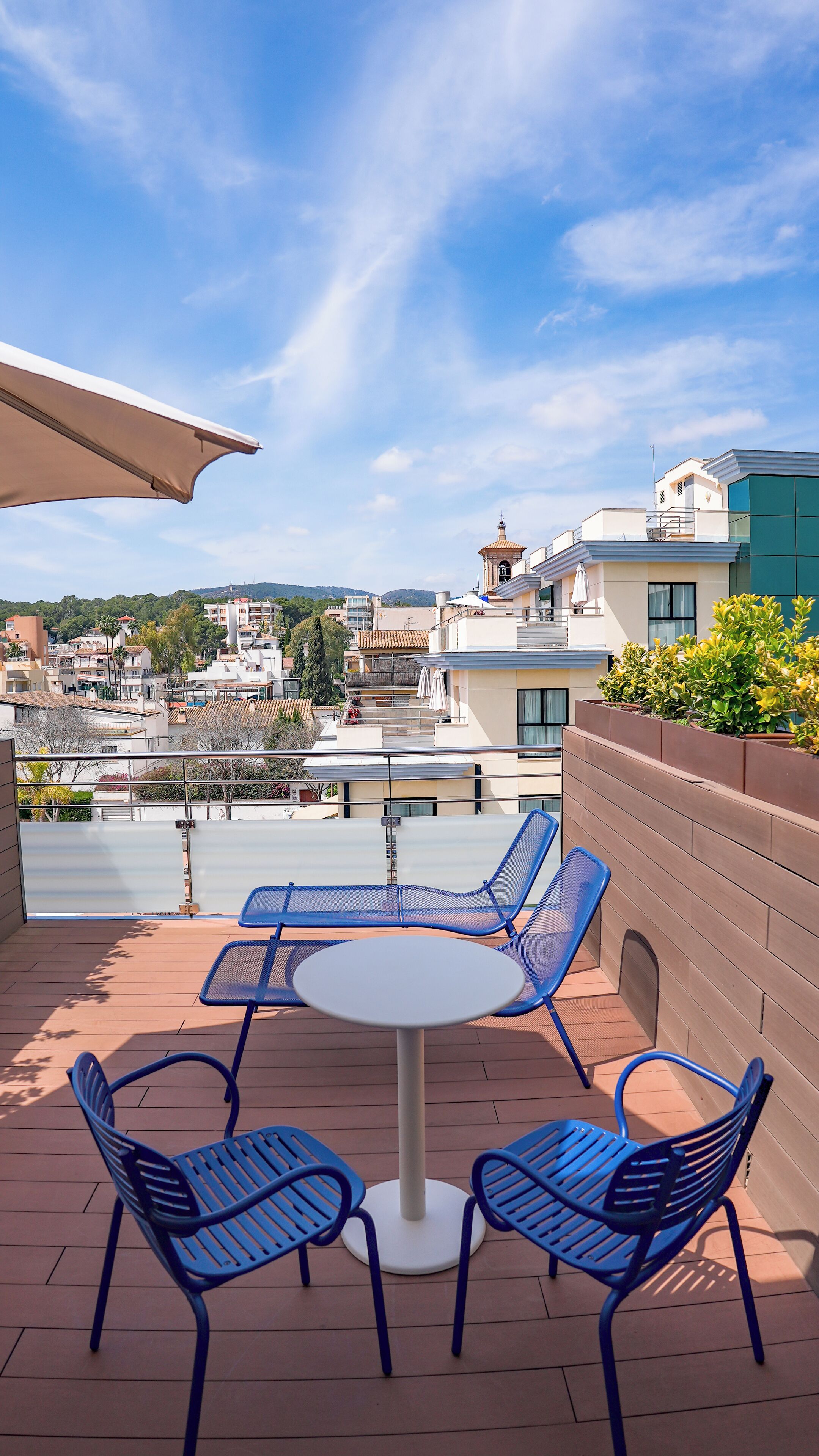 A rooftop terrace with a round table and four blue chairs, overlooking a cityscape with buildings and a hill in the distance.
