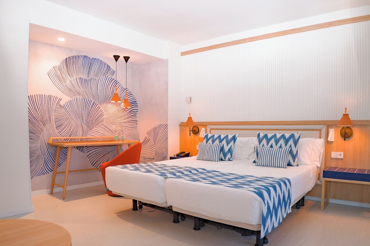 Une chambre d’hôtel moderne avec un grand lit, un bureau et une chaise. Un mur présente un motif original, et une grande fenêtre est dotée de stores.