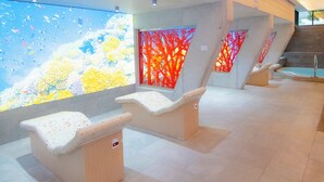 A modern spa area with three wicker loungers, a large digital screen displaying a nature scene, and a red abstract wall. - Hotel Isla Mallorca & Spa (Palma de Mallorca)