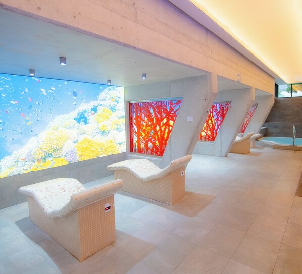 Un área de spa moderna con tres sillones de mimbre, una amplia pantalla digital que muestra un paisaje natural y una pared abstracta roja.