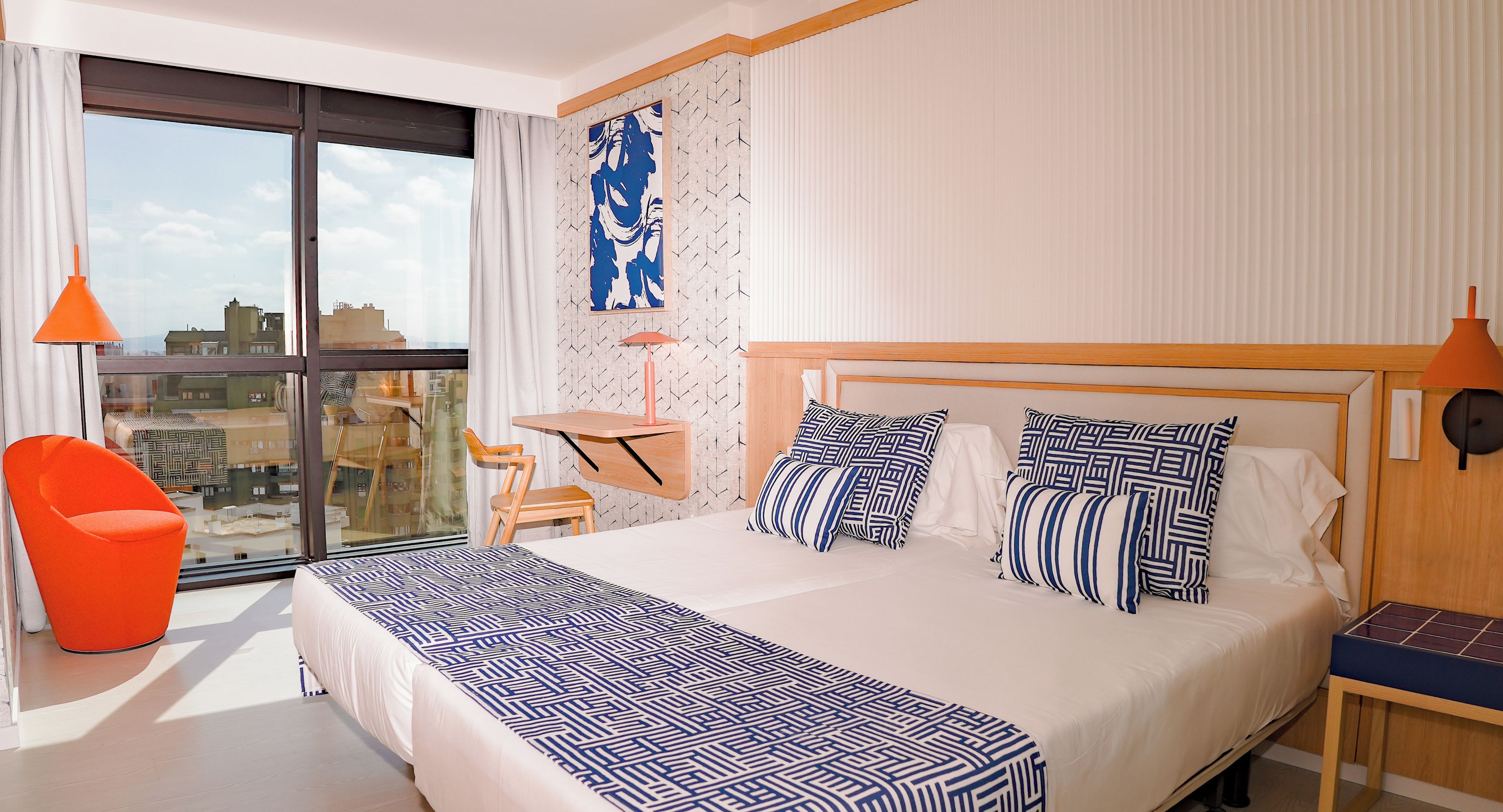 Ein Hotelzimmer mit einem großen Bett, einem Schreibtisch, einem Stuhl und Blick auf die Stadt durch ein großes Fenster.