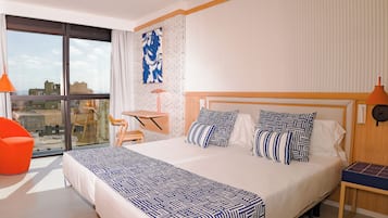 Habitación de hotel con una cama grande, un escritorio, una silla y vistas de la ciudad a través de un ventanal amplio.