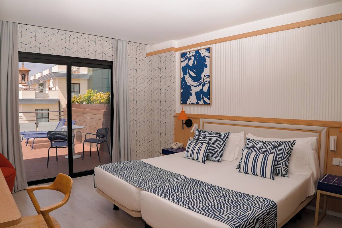 Une chambre d’hôtel comprenant un lit, un bureau, une chaise, un balcon avec une table et des chaises, et une vue sur les bâtiments environnants.