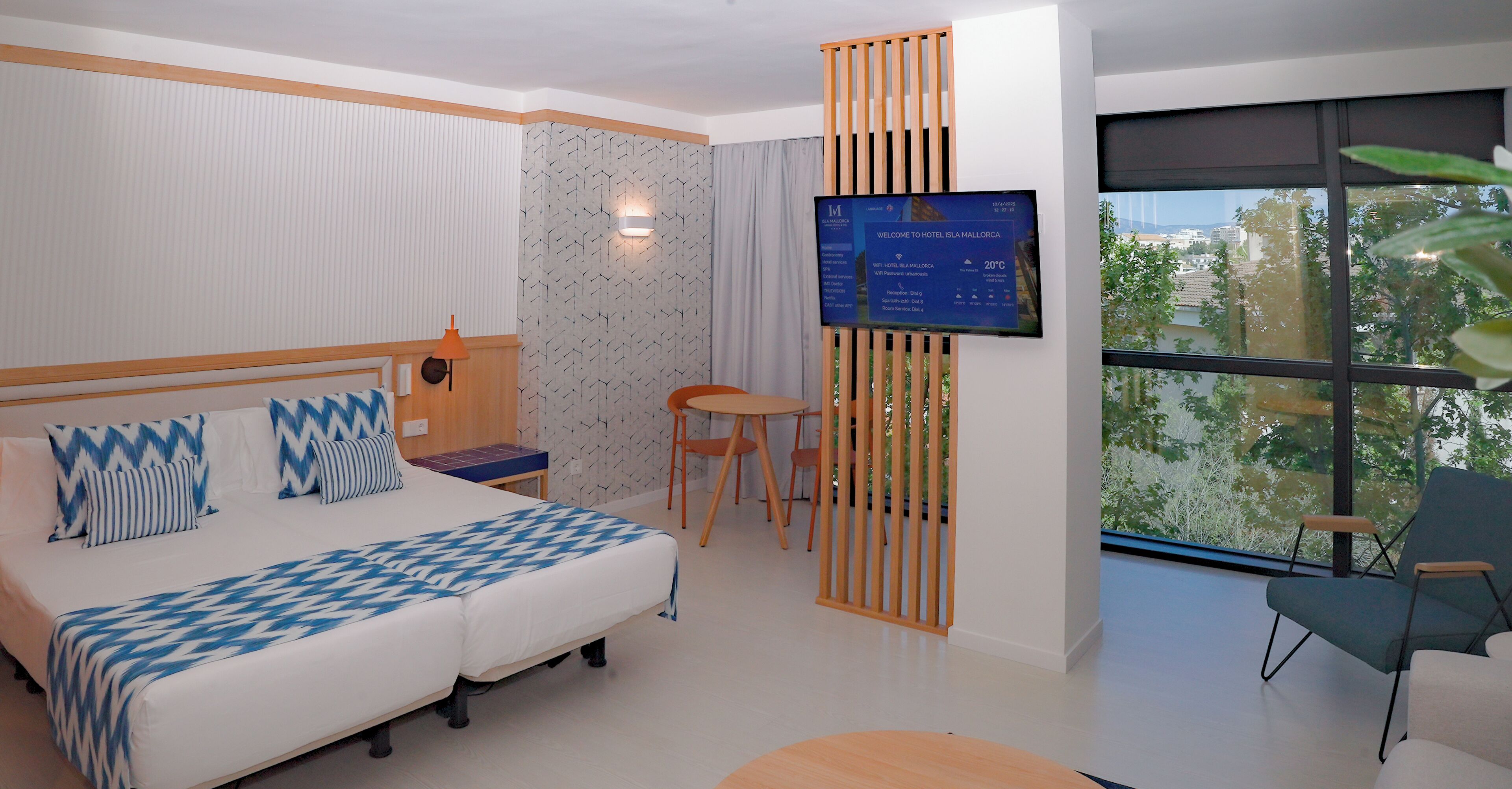 A modern hotel room with a bed, a desk, a chair, and a TV displaying room information.