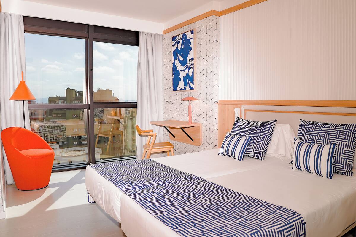 Une chambre d’hôtel avec un grand lit, un bureau, une chaise et une vue sur la ville grâce à une grande fenêtre.