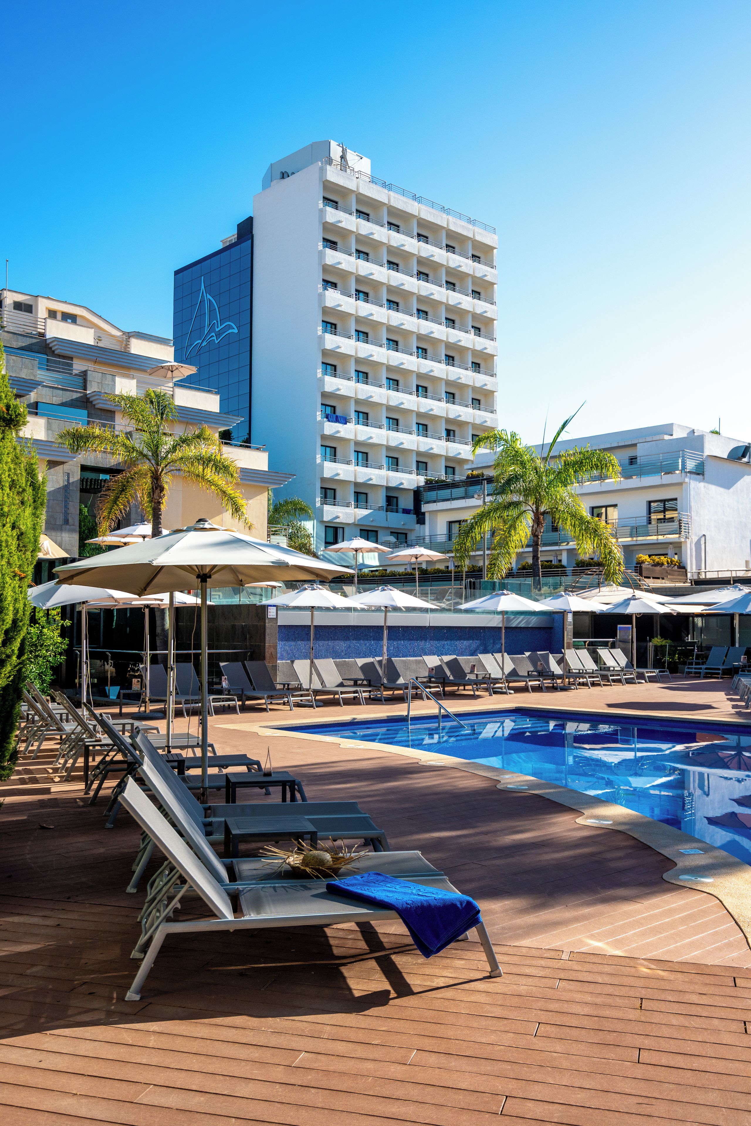 A hotel pool area with lounge chairs and umbrellas, a clear blue sky, and a tall building in the background.