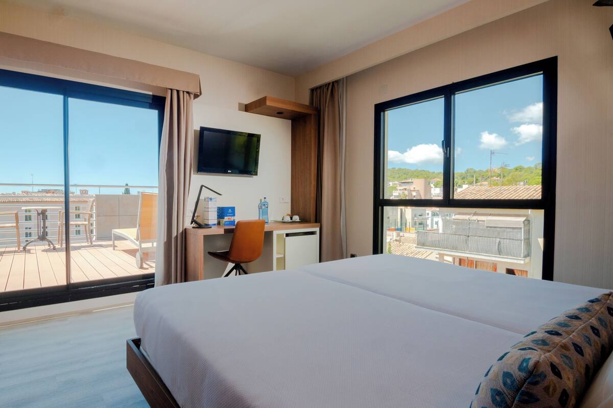 Une chambre d’hôtel dotée d’un grand lit, d’un bureau avec une chaise, d’une télévision et de grandes fenêtres offrant une vue sur les bâtiments et un ciel dégagé.