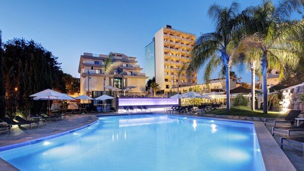 A swimming pool with lounge chairs and umbrellas, surrounded by palm trees and a hotel building. - Hotel Isla Mallorca & Spa (Palma de Mallorca)