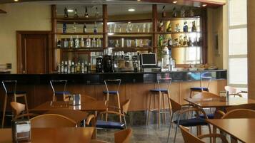 Zona de bar con sillas y mesas de madera, una barra con diferentes botellas y vasos, y una máquina de café.