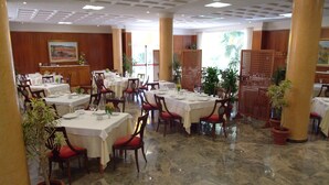 Un área de comedor con varias mesas y sillas, algunas con cojines rojos, y plantas en macetas alrededor.