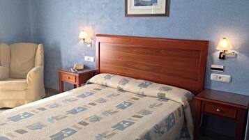 Un dormitorio con cabecera de madera, cama con edredón estampado, dos mesitas de noche con lámparas, una silla e imagen enmarcada en la pared.