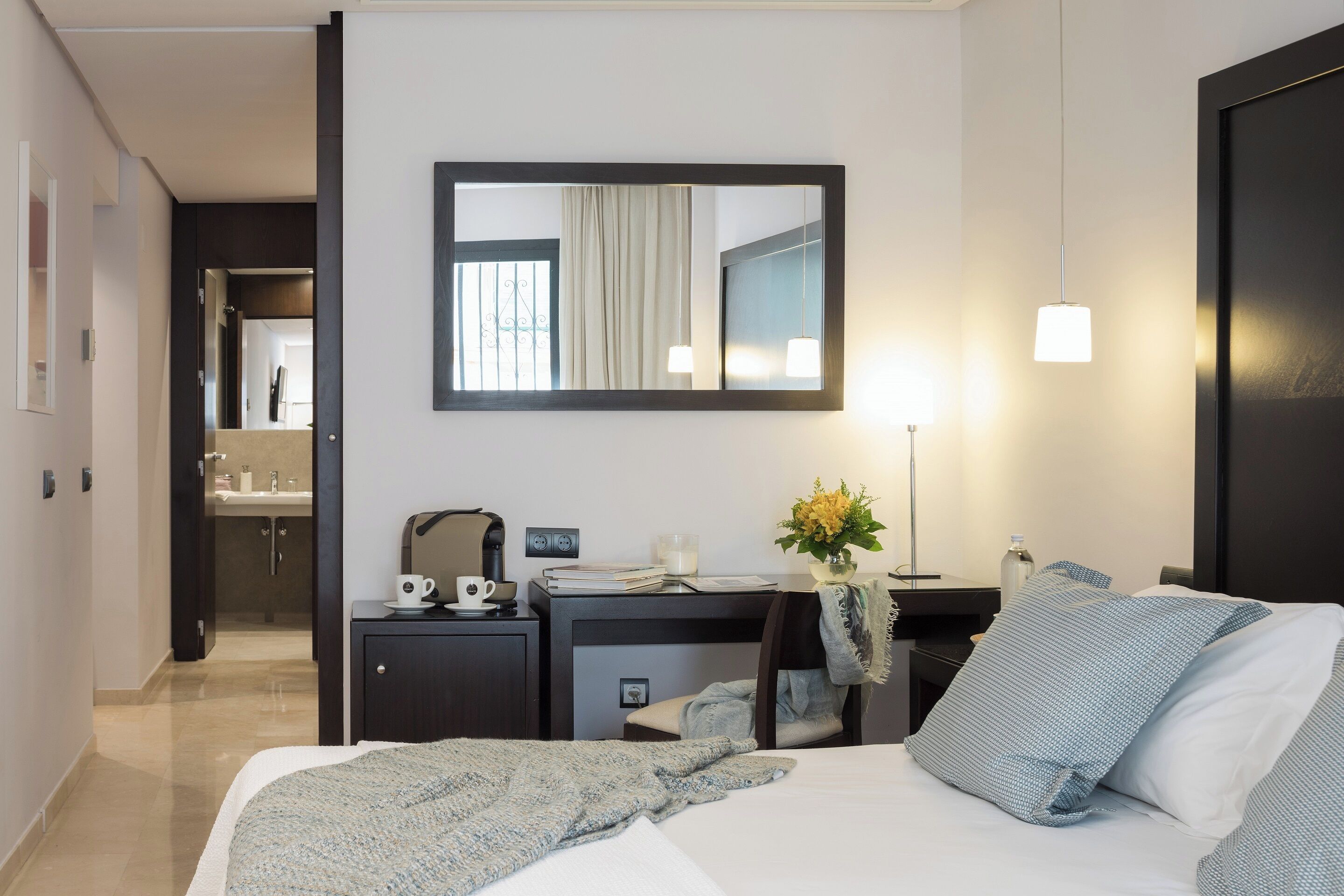 A hotel room with a bed, a desk with a computer, a mirror, and a nightstand with a lamp and a vase of flowers.